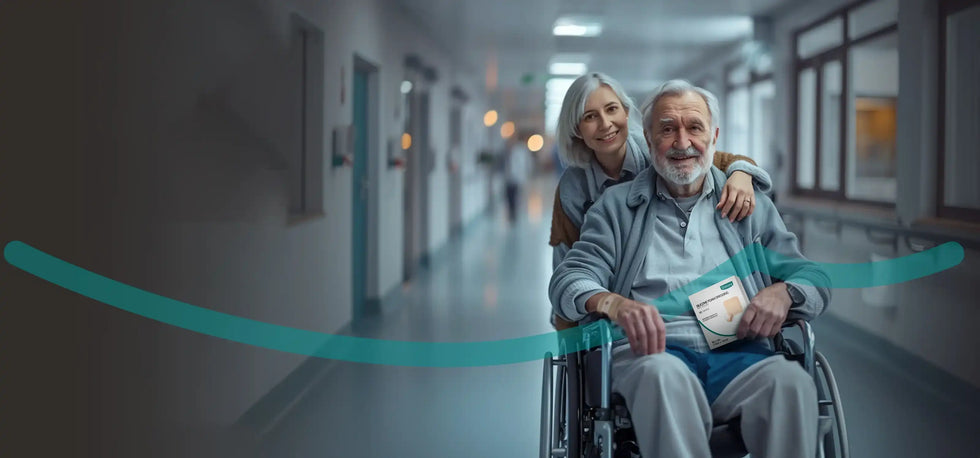 Smiling elderly man in wheelchair holding medical wound care product, with caregiver in hospital corridor